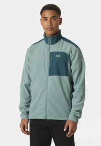 Chaqueta de forro polar verde claro con cremallera frontal, cuello alto y paneles de acento en azul marino; cuenta con un bolsillo en el pecho y mangas largas.