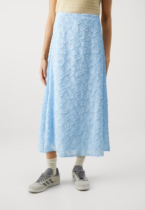 Fustă bleu deschis, de lungime medie, cu broderie florală, talie bine definită și siluetă fluidă. Purtați cu sneakers gri și șosete ușoare.