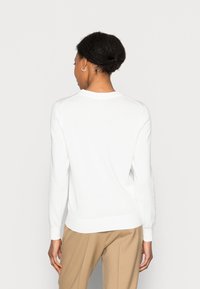 Maglione bianco a maniche lunghe con scollo rotondo e polsini a costine, abbinato a pantaloni beige per il contrasto.