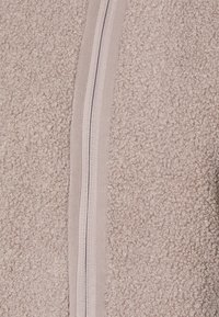 Ljus beige fleecejacka med en texturerad yta, som har en slät dragkedja framtill och en kontrasterande kant längs dragkedjan.