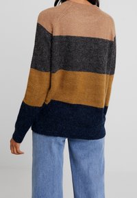 Knit sweater with horizontal stripes in beige, dark gray, mustard yellow, and navy. Features a round neck and loose fit, paired with light denim.