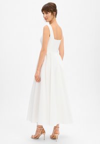 Robe blanche sans manches avec un dos carré, une jupe évasée et une texture lisse. Portée avec des talons beiges à brides pour un look complet.
