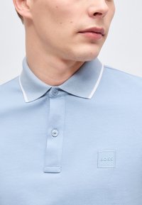 Homme portant un polo bleu clair avec un bord blanc au niveau du col et deux boutons, arborant un petit logo "BOSS" embossé sur la poitrine.