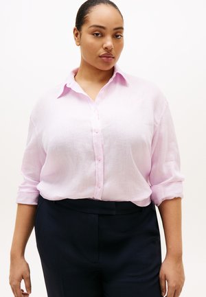 Femeie purtând o cămașă roz deschis cu nasturi și mâneci suflecate și pantaloni închiși la culoare, stând în fața unui fundal alb simplu.