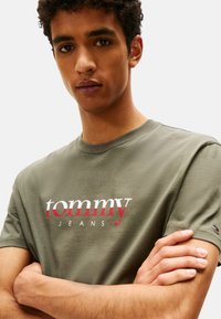 T-shirt in cotone verde oliva con logo "tommy JEANS" rosso e bianco centrato, maniche corte, colletto alla giapponese e un piccolo dettaglio del logo sulla manica.