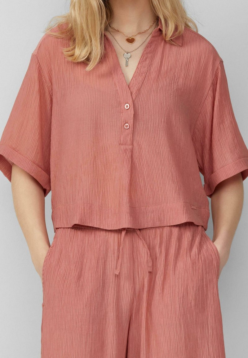 Haut court à manches courtes avec boutons, en tissu doux, texturé et rose poussiéreuse. Il présente un col en V et de fines côtes verticales discrètes.