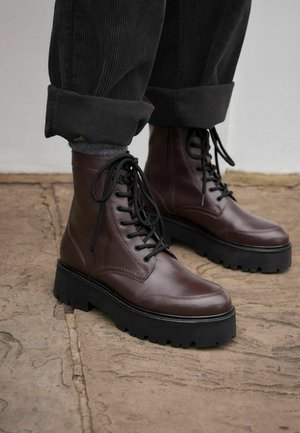 Brown leather ankle boots with black laces, a rounded toe, and a thick, ridged black sole, worn with folded black corduroy pants.