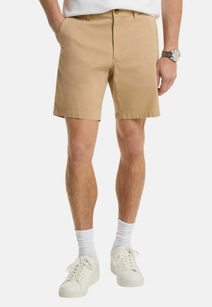 Uomo che indossa pantaloncini beige, calzini bianchi, scarpe da ginnastica bianche e un orologio, in piedi con le mani parzialmente in tasca contro uno sfondo semplice.