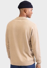 Pull en maille beige clair avec un motif en nid d'abeille texturé, col et poignets côtelés. Associé à un pantalon en denim foncé et un beanie marine.