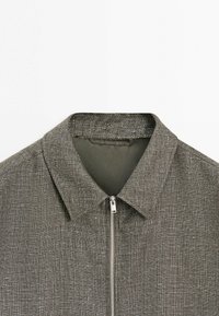 Grå tekstureret herrejakke med en spids krave og en sølvfarvet lynlås foran, vist mod en hvid baggrund.