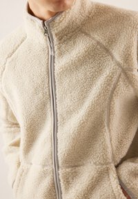 Cream-colored fleece jacket with a high collar, zip closure, and textured, fluffy surface. Features contrasting gray stitching accents.