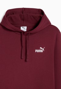 Burgundska dukserica od mekog materijala s kenguruvim džepom. Ima bijeli Puma logo na prsima i kapuljaču s vezicom.
