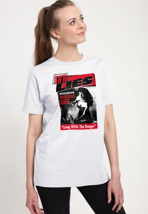 Frau mit braunen Haaren im Pferdeschwanz, die ein weißes T-Shirt mit rotem und schwarzem Text und einer schwarz-weißen Grafik trägt.