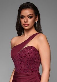 Robe de soirée bourgogne à une épaule, dotée d'un corsage orné de sequins et d'un tissu lisse drapé. Accentuée d'éléments transparents.