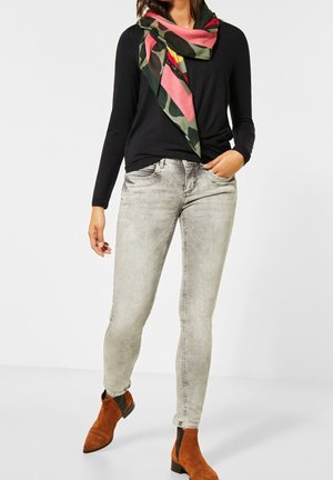 Kobieta w czarnej bluzce z długim rękawem, wyblakłych szarych dopasowanych dżinsach, brązowych botkach nad kostkę oraz wielokolorowym szaliku z różowymi, żółtymi i zielonymi wzorami.