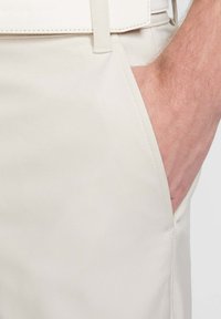 Pantalones beige a medida con una textura de tejido suave, bolsillos laterales y detalles cosidos. Trabillas en la cintura, con un ligero brillo.