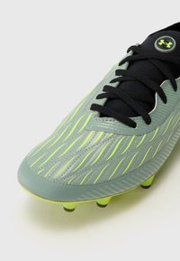 Scarpino da calcio verde lime e grigio con una superficie testurizzata, strisce sottili e lacci neri. Presenta un logo nero sulla linguetta e tacchetti sagomati.