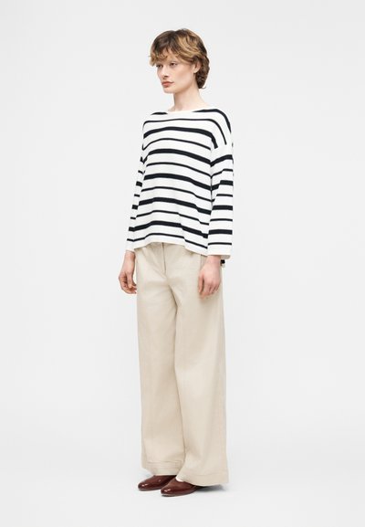 Άτομο που φοράει λευκό μπλουζάκι μάκρους μανικιού με μαύρες οριζόντιες ρίγες, μπεζ παντελόνι με φαρδιά πόδια και καφέ παπούτσια, στέκεται μπροστά σε λευκό φόντο.