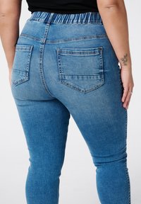 Vervaagde blauwe denim jeans met een elastische tailleband, voorzien van twee achterzakken en een slim fit ontwerp. Subtiele wassing nabij de tailleband.