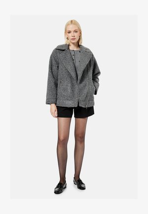 Lyshåret kvinde iført en grå oversized jakke med struktur, grå top, sorte shorts, gennemsigtige sorte strømpebukser og sorte loafers, stående mod en hvid baggrund.