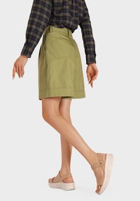Pantaloni corti verde chiaro con tasche laterali, abbinati a sandali con platform color cammello dotati di cinghie incrociate. La parte superiore è una camicia a quadri blu navy e verde.