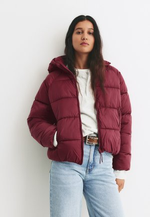 Burgunderfarvet pufferjakke med hætte, der har lynlås fortil og sidelommer. Tekstureret, quiltet design kombineret med lyseblå jeans.