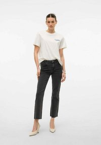 Camiseta blanca de algodón con gráfico azul, combinada con jeans negros ajustados de cintura alta y tacones en color crema. Un atuendo simple y causal.