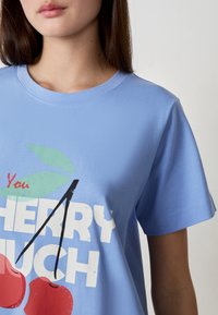 T-shirt di cotone azzurro chiaro con una grafica che presenta ciliegie rosse, foglie verdi e la frase "You Cherry Much" in caratteri bianchi in grassetto.