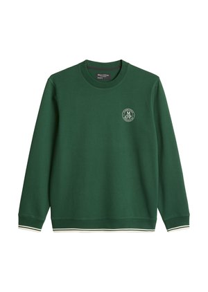 Jeune homme portant un sweat-shirt vert et un short blanc, tenant un sac de sport vert, debout devant un fond blanc uni.