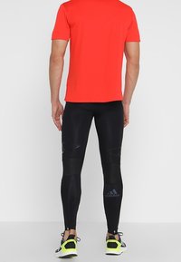 Maglietta atletica rossa a maniche corte abbinata a leggings neri aderenti con dettagli in mesh e zip alle caviglie, indossata con scarpe nere e gialle.