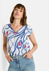 Top de manga corta en tejido ligero con un patrón abstracto en azul, blanco y rojo, con un acabado texturizado. Lleva puestos unos vaqueros.