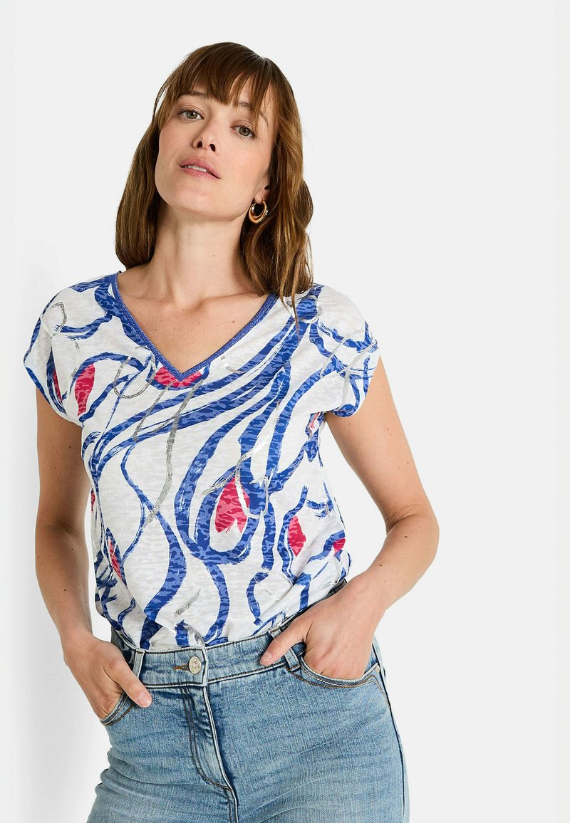 Top de manga corta en tejido ligero con un patrón abstracto en azul, blanco y rojo, con un acabado texturizado. Lleva puestos unos vaqueros.