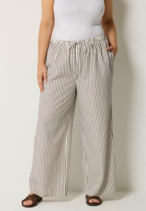 Femme portant un pantalon à jambes larges à rayures verticales blanches et marron avec une taille élastique et un cordon de serrage, associée à des sandales marron et un haut blanc.