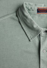 Polo verde chiaro con colletto, apertura con bottoni, cuciture visibili e etichetta Superdry all'interno della scollatura.