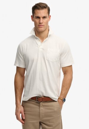Uomo con capelli corti che indossa una polo bianca con colletto alzato, pantaloni beige, cintura di pelle marrone e orologio da polso con quadrante blu.