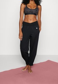 Žena stojící bosky na růžové cvičební podložce, oblečená v černém vzorovaném sportovním podprsence a černých teplácích Adidas, s rukama v kapsách.