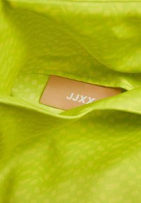 Forro interior verde brillante con un patrón geométrico, que incluye una etiqueta marrón con "JJXY" en texto blanco. Material suave y texturizado.