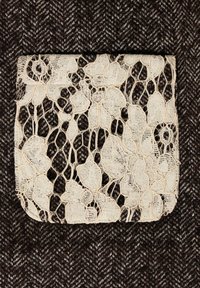 Krémová krajková kapsa s květinovým vzorem na tmavě hnědém rybí kosti tkaném pozadí, ukazující složité detaily a strukturu.