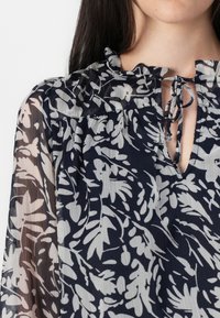 Blusa a fiori in bianco e nero con colletto arricciato e dettaglio con fiocco, indossata da un modello. Il tessuto sembra leggero e trasparente.