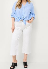 Camicetta azzurra chiara con collo a bottoni e nodo frontale, maniche arrotolate; abbinata a pantaloni bianchi a gamba larga. Sandali con tacco a blocco aperti neri completano l'outfit.
