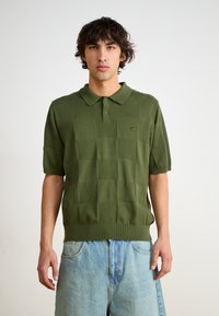 Grønn strikket polo med kort erm, med et teksturert rutemønster og ribbestrikket kant, kombinert med lyseblå denimshorts.