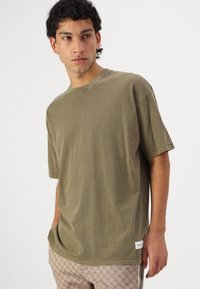Oliven grøn bomulds t-shirt med en afslappet pasform, rund halsudskæring, korte ærmer og diskrete syninger, bæres med mønstrede beige bukser.