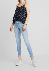 Femme portant un jean skinny bleu clair roulé aux chevilles, des baskets blanches et un haut sans manches à motifs paisley noir.