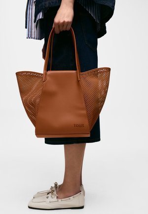 Persona sosteniendo un gran bolso de cuero marrón con laterales perforados, vistiendo una falda oscura y zapatos blancos tipo slip-on con lazos.