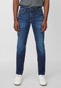 Jeans de mezclilla de lavado oscuro con ajuste slim, que presentan un ligero desgaste, cinco bolsillos y cierre de botón. Combinados con zapatillas blancas.