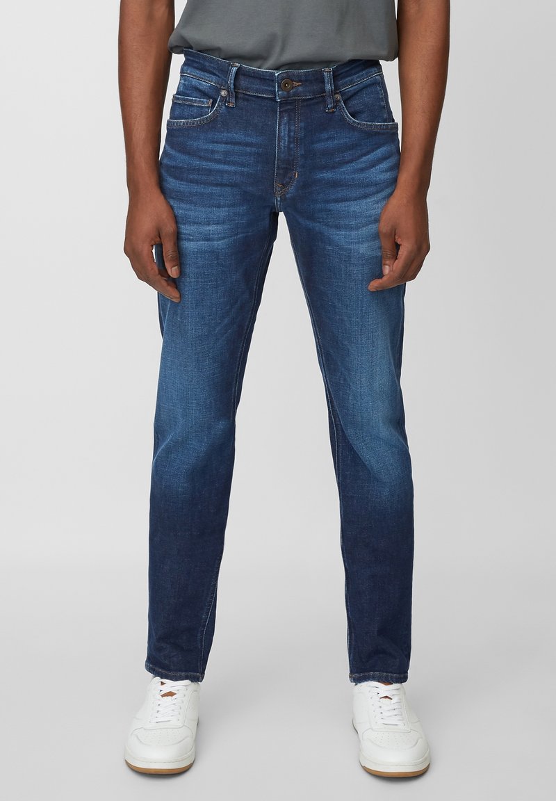 Jeans de mezclilla de lavado oscuro con ajuste slim, que presentan un ligero desgaste, cinco bolsillos y cierre de botón. Combinados con zapatillas blancas.