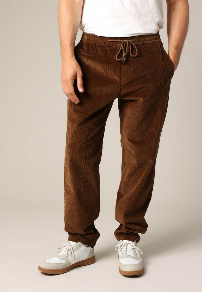 Pantalon en velours côtelé marron à coupe décontractée, doté d'une taille élastique et d'un cordon de serrage. Associé à des baskets blanches et grises.