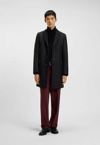 Schwarzer Wollmantel mit Reverskragen, sechs Knöpfen und Seitentaschen; getragen über einem schwarzen Rollkragenpullover und bordeauxfarbenen Hosen, accessorized mit schwarzen Schuhen.