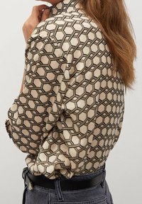 Cremefarbene Satinbluse mit einem geometrischen hexagonalen Muster in Schwarz. Sie hat einen hohen Ausschnitt und weit geschnittene Ärmel, kombiniert mit Jeans.