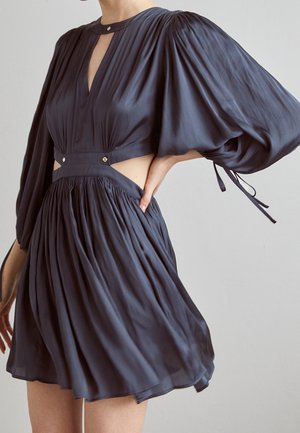 Femme portant une robe bleu foncé avec de longues manches bouffantes, des découpes sur les côtés de la taille, une jupe plissée et de petits détails de boutons métalliques.